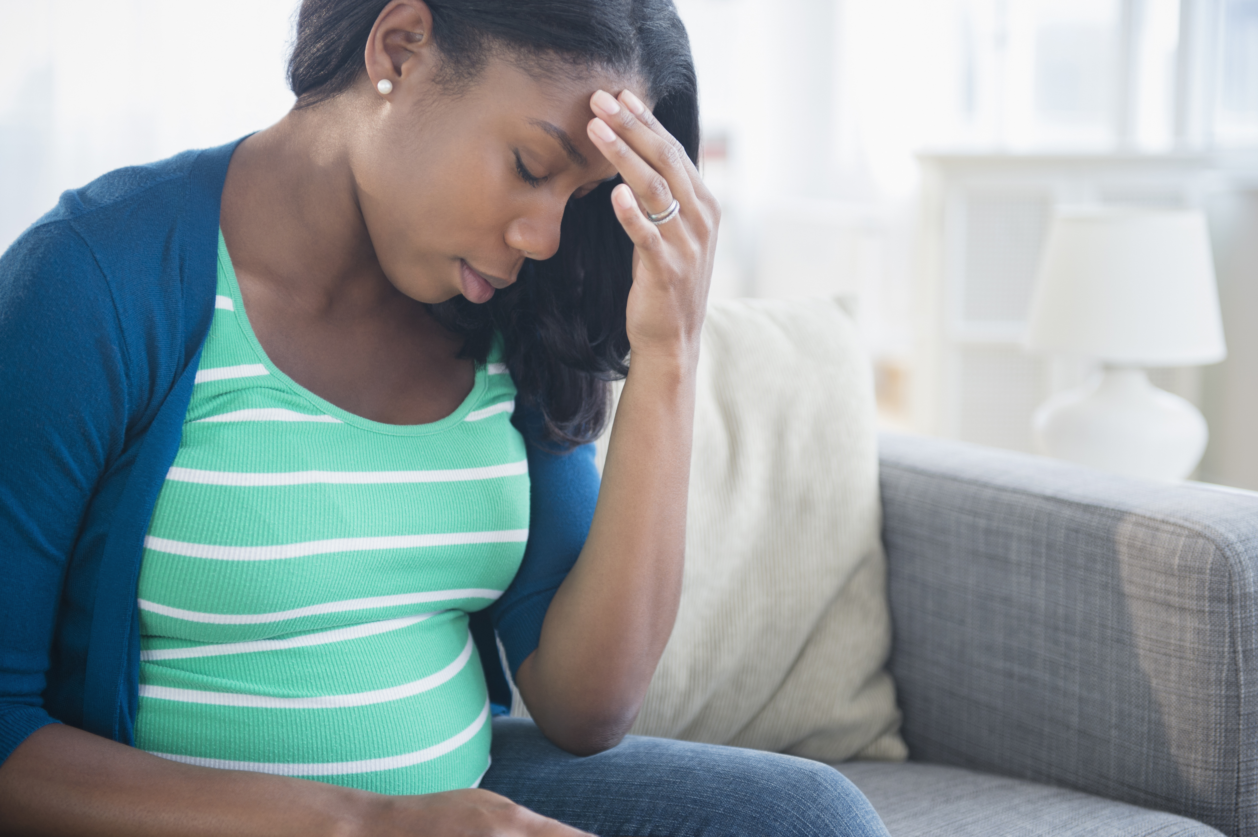 Anxious Black pregnant woman rubbing forehead on sofa.